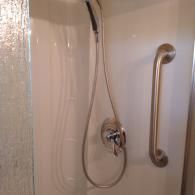 Shower stall interior with a handheld shower head, a chrome mixing valve, and a vertical brushed nickel grab bar.