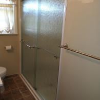 A view of a bathroom shower featuring textured glass sliding doors, chrome towel bars, and a nearby toilet and window.