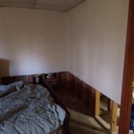 A bedroom under renovation showing a partially removed wall with exposed wooden framing studs and wood paneling base.