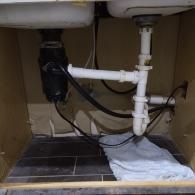 Under-sink area showing a garbage disposal connected to white PVC drainage pipes and a folded white towel on the floor.