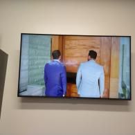 Two men in suits standing side-by-side in front of a wooden door on a television screen.