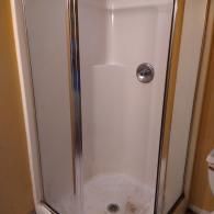 A corner shower stall with white walls, a glass enclosure, and a silver handle in a room with tan walls.