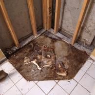 An unfinished bathroom corner with exposed wall studs and a recessed, debris-covered concrete floor around a drain.