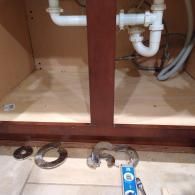 The interior of a wooden cabinet under a sink with white plumbing pipes, tools, and metal hardware on the tiled floor.
