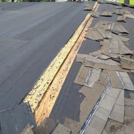 A roof under renovation, showing a wooden ridge board separating smooth underlayment and torn asphalt shingles.