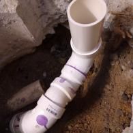 A white PVC pipe assembly with multiple elbows and couplings installed in a dirt trench next to a concrete wall.