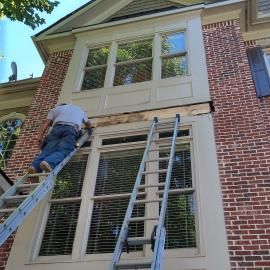 A person on a ladder repairs the exterior wood trim between two stacked windows on a brick house.