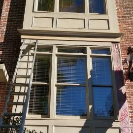 A person on a ladder repairs exterior trim around a large, multi-paned window on a red brick house.