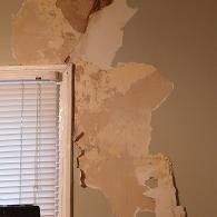 A white window blind next to a wall with large patches of drywall paint peeling off to reveal the brown plaster underneath.
