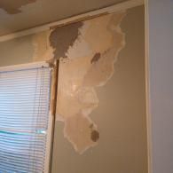 A damaged indoor wall with peeling paint and exposed plaster near a window with white blinds.