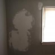A wall with large, irregular patches of light-colored drywall compound over dark gray paint near a window.