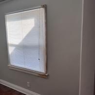 A window with white horizontal blinds is centered on a light gray wall with white trim.