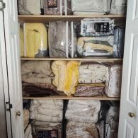 A closet organized with four shelves full of neatly stacked, packaged blankets and bedding in various colors and textures.