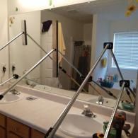 A large bathroom mirror being held in place by two adjustable metal support poles during installation over a double vanity.