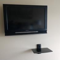 A black television mounted on a beige wall, positioned above a small, empty black glass wall-mounted shelf.
