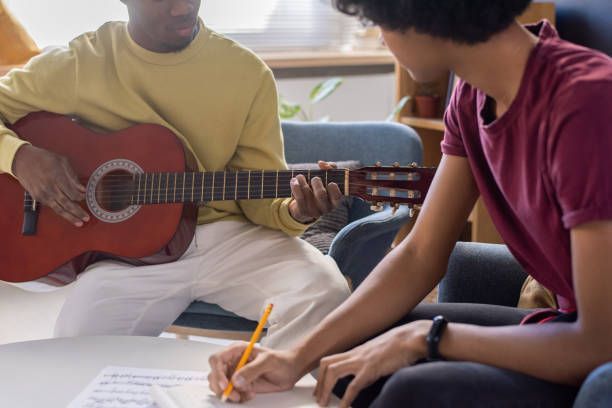 Young guy taking Guitar Lessons in Holly Springs, NC, noting music as teacher plays guitar at home.