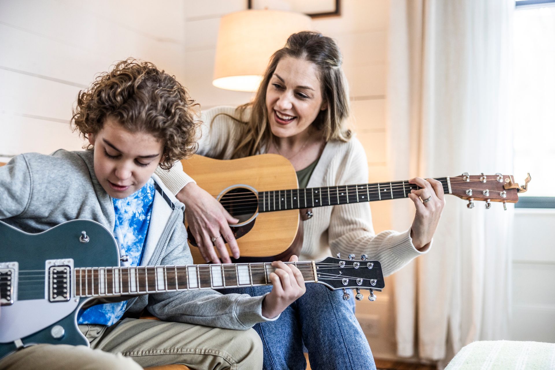 Smiling woman teaching a boy electric and acoustic guitar music lessons in a cozy room. 