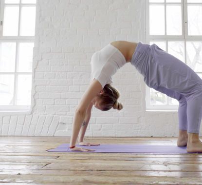 A person in a white top and purple pants performing a bridge yoga pose on a mat in a white, sunlit room with wooden floors.
