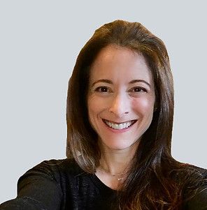 A smiling person with shoulder-length brown hair wearing a black top against a plain light gray background.