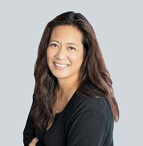 A smiling person with long brown hair, wearing a black shirt, posing against a solid light gray background.