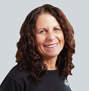 A smiling person with shoulder-length, wavy dark hair, wearing a black shirt against a light gray background.
