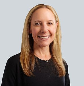 A smiling woman with shoulder-length blonde hair, wearing a black top and a delicate necklace