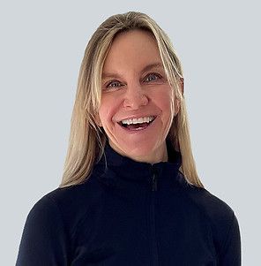 A smiling person with long blonde hair wearing a dark, high-necked zip-up top against a plain light gray background.
