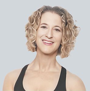 A person with curly shoulder-length hair smiles warmly against a light grey background, wearing a black athletic top.