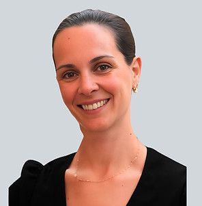 Headshot of a smiling person with dark hair pulled back, wearing a black top and a thin gold necklace against a gray wall.