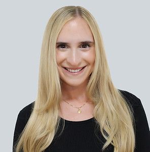 A smiling person with long blonde hair, wearing a black shirt and a simple gold necklace against a plain gray background.