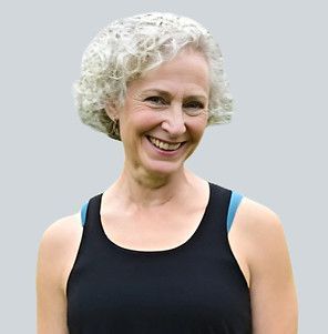 A smiling person with curly gray hair, wearing a black tank top, set against a plain light gray background.
