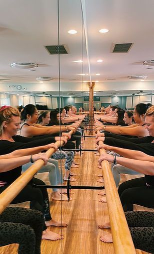 People hold onto a wooden ballet barre during a fitness class in a room with a large mirror reflecting the group.
