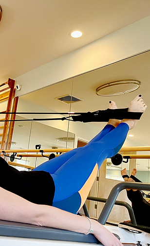 A person in blue leggings performs a Pilates exercise with their feet in straps while lying on a reformer machine.