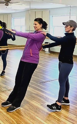 An instructor in a cap assists a participant with a standing back-extension exercise in a dance studio with mirrors.