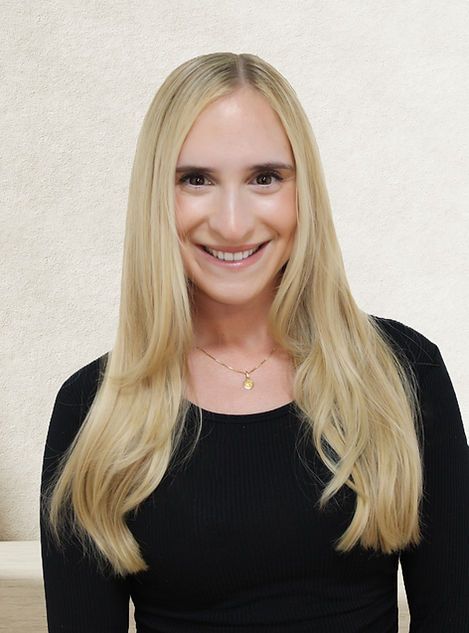 A person with long blonde hair, smiling, wearing a black long-sleeved shirt and a gold pendant necklace against a beige wall.