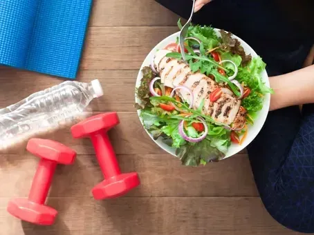 A plate of grilled chicken salad, two red dumbbells, a water bottle, and a yoga mat on a wooden floor.