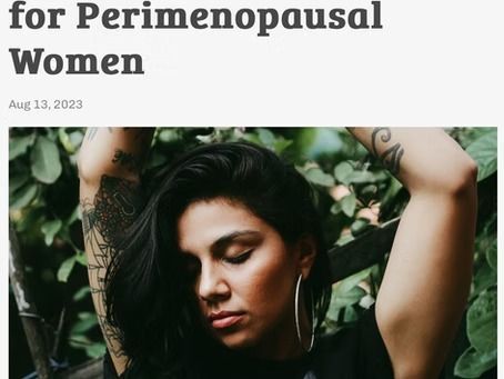 A person with tattoos on both arms poses with their hands behind their head amidst green foliage.