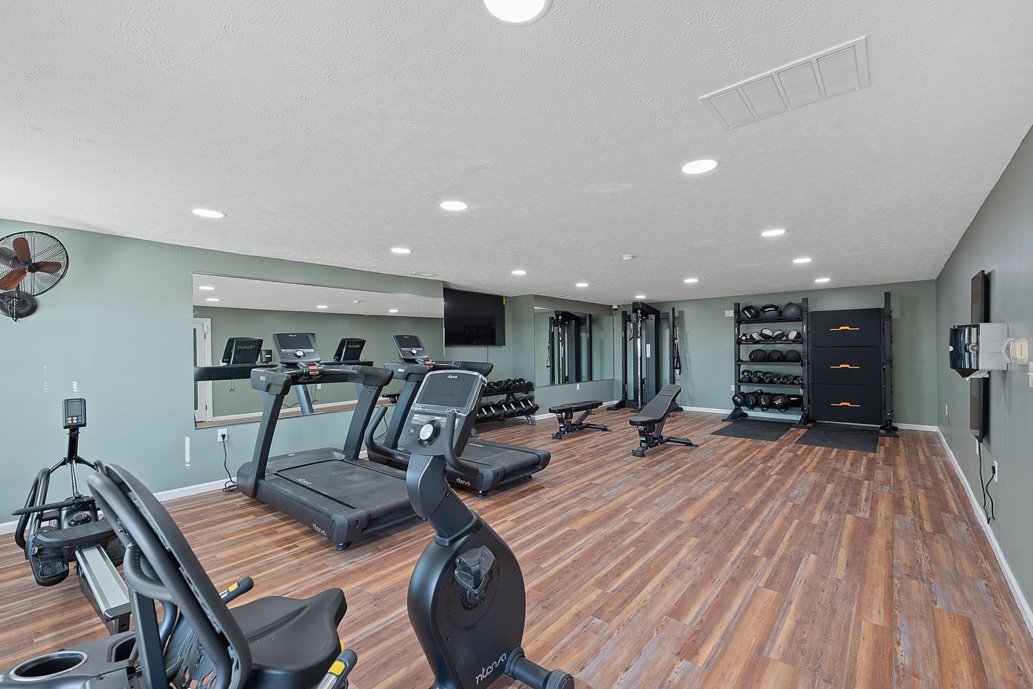 Gym interior with cardio equipment and weights on a wood floor. Mirrors line a wall, and a TV is mounted.