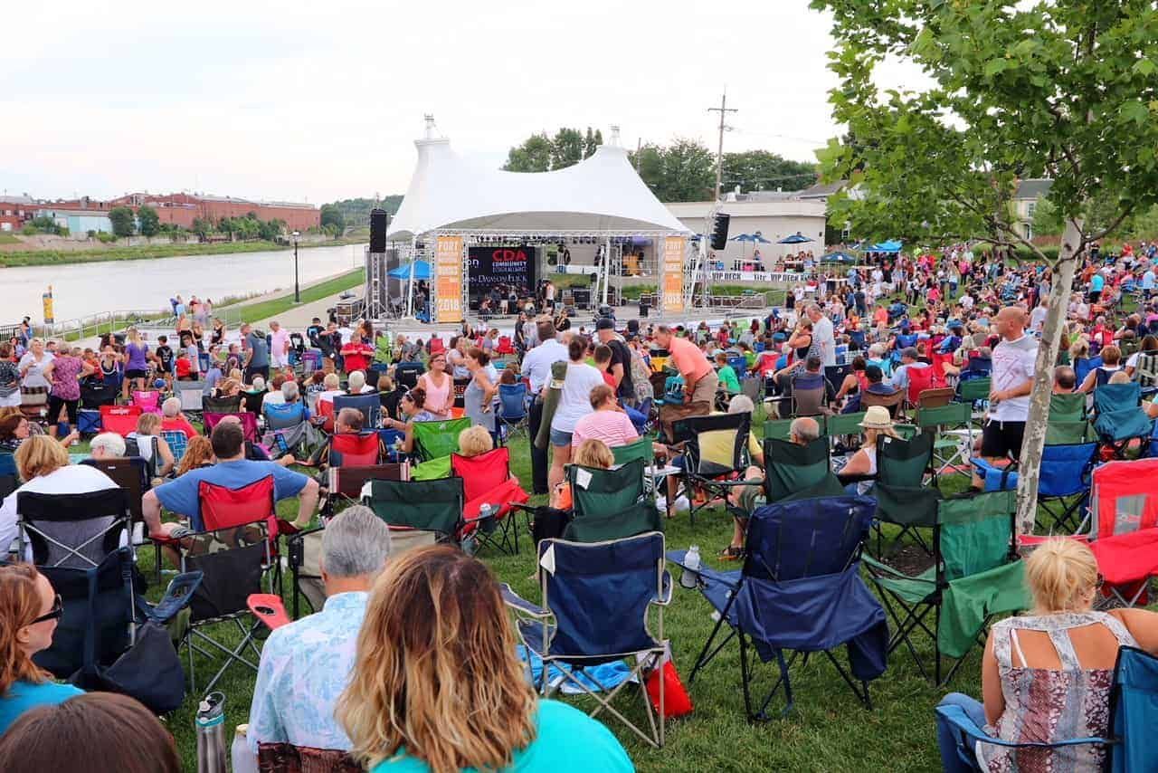 Large crowd at outdoor concert, stage with band, green grass, river, buildings in background.