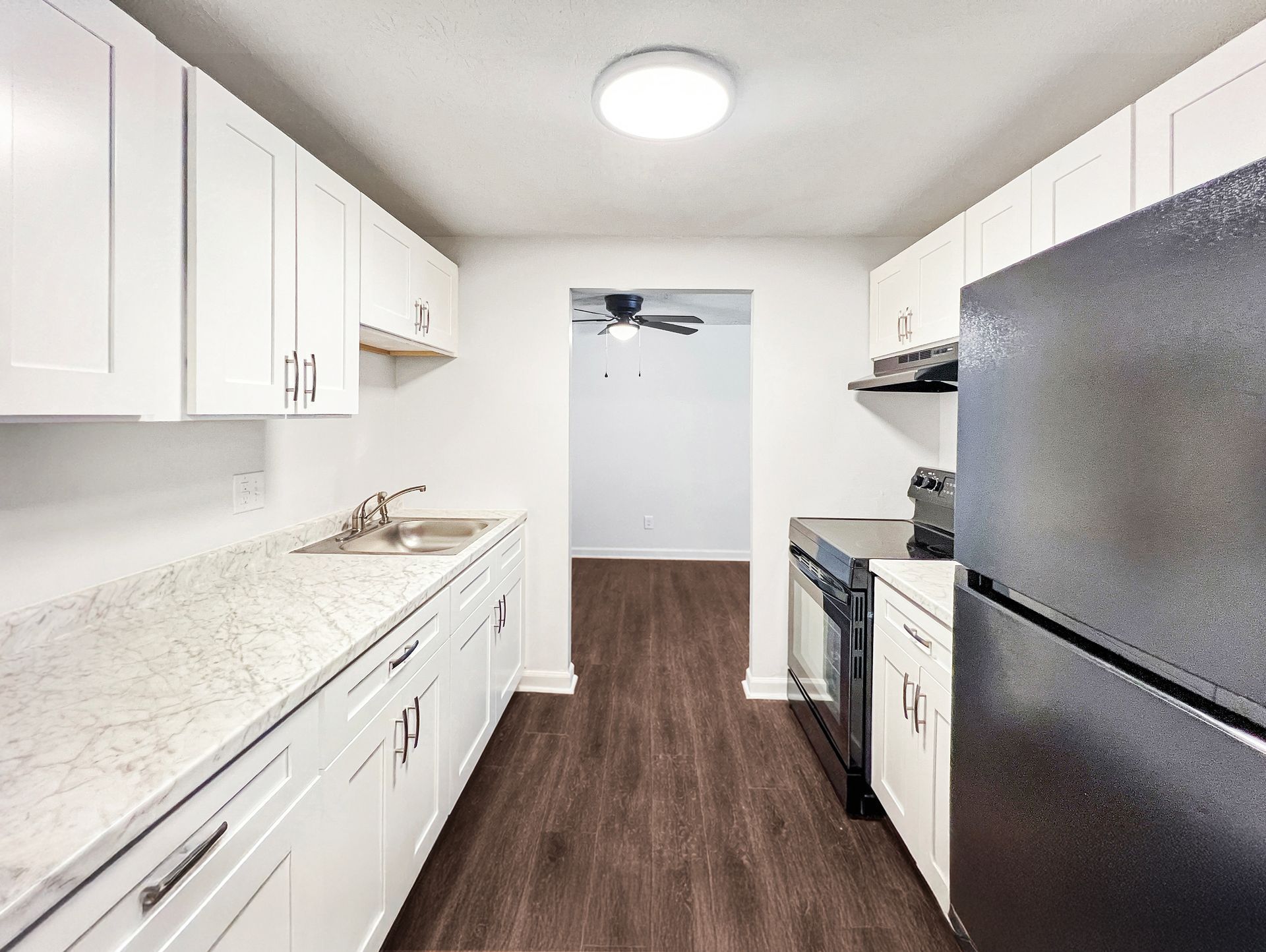 White kitchen with black appliances, leading to a doorway and dark wood-look flooring.