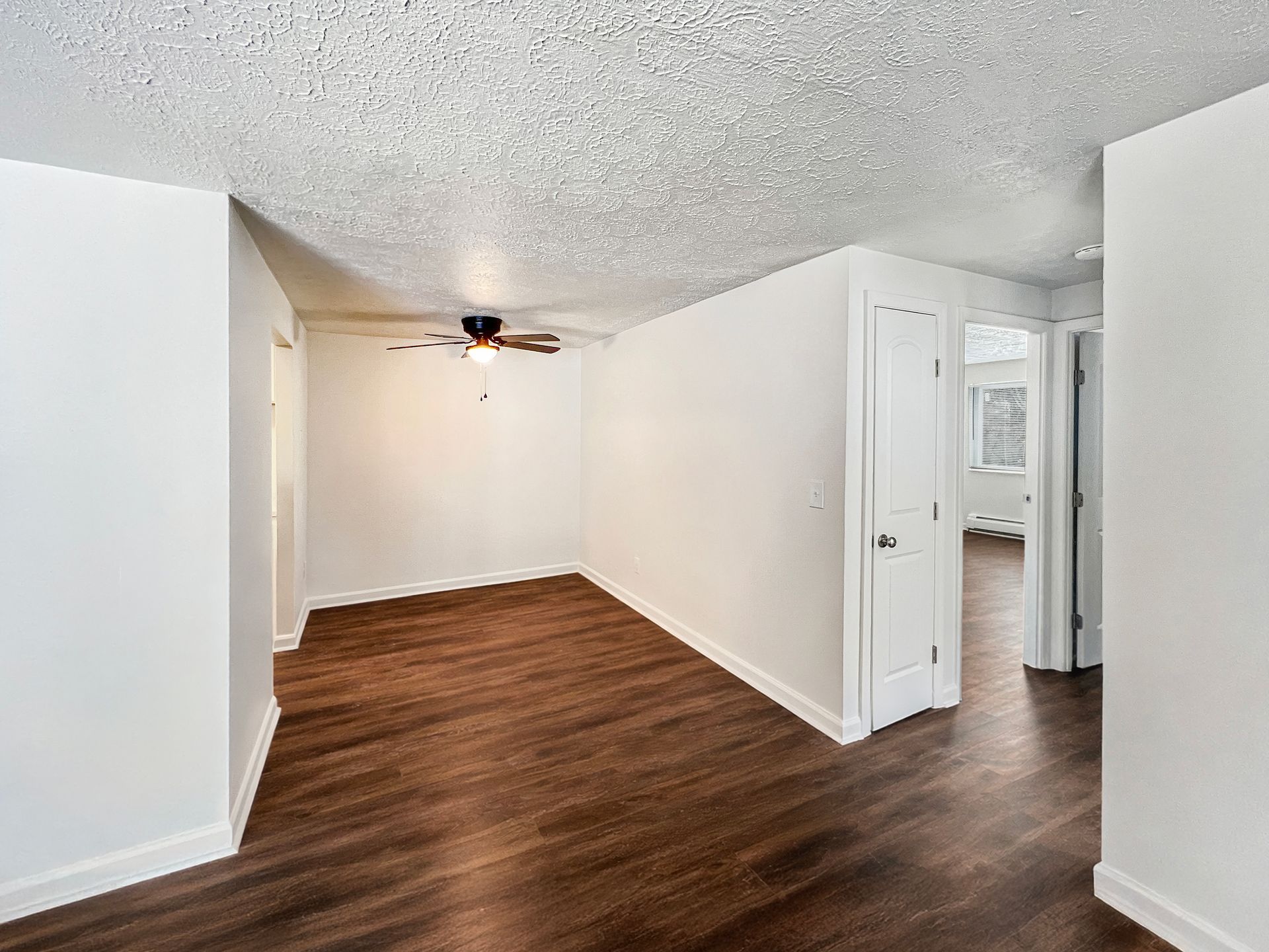 Empty room with dark wood flooring, white walls, and a popcorn ceiling. A door and an open doorway are visible.
