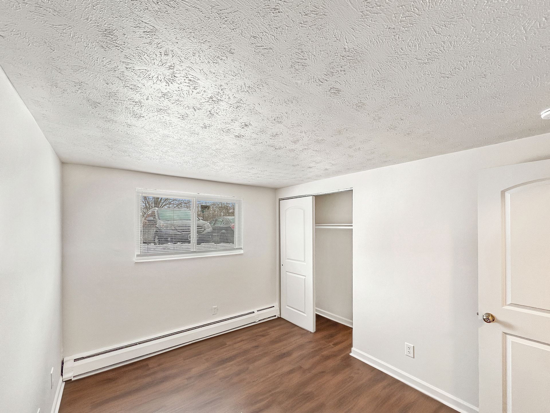 Empty bedroom with wooden floor, white walls, closet, window, and textured ceiling.