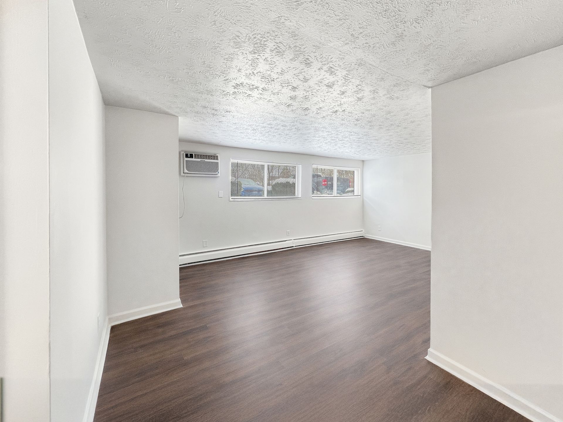 Empty, bright room with dark wood-look flooring, white walls, and three windows.