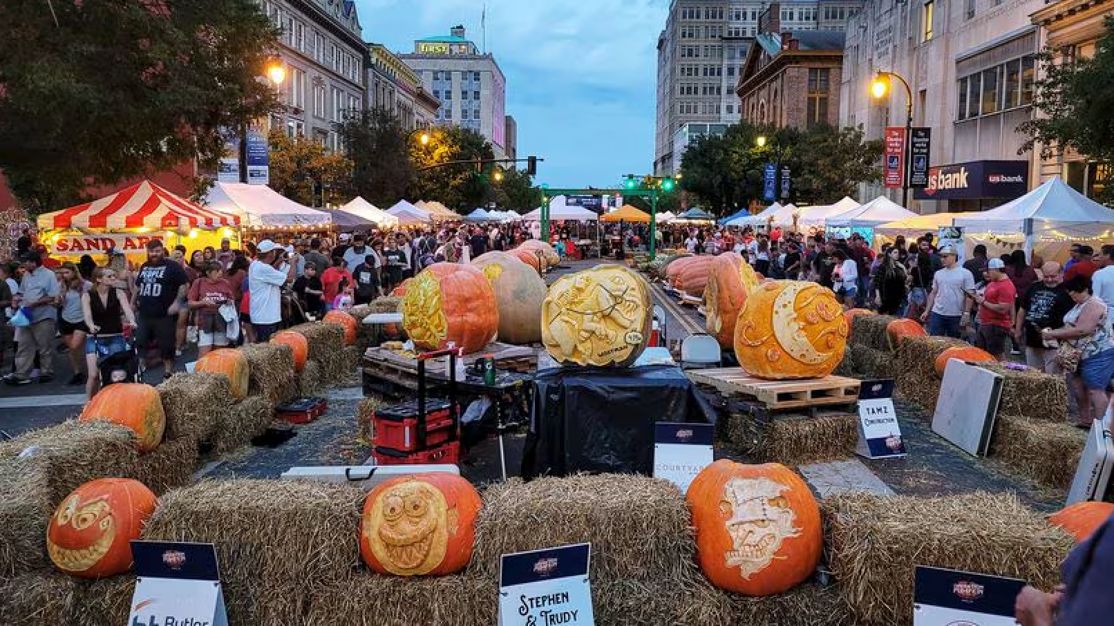 Street festival with carved pumpkins on display; people walking, white tents, buildings in background.