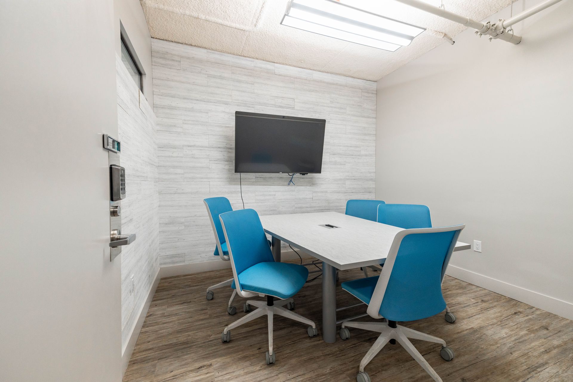 Small conference room with a wall-mounted TV, white table, and blue office chairs