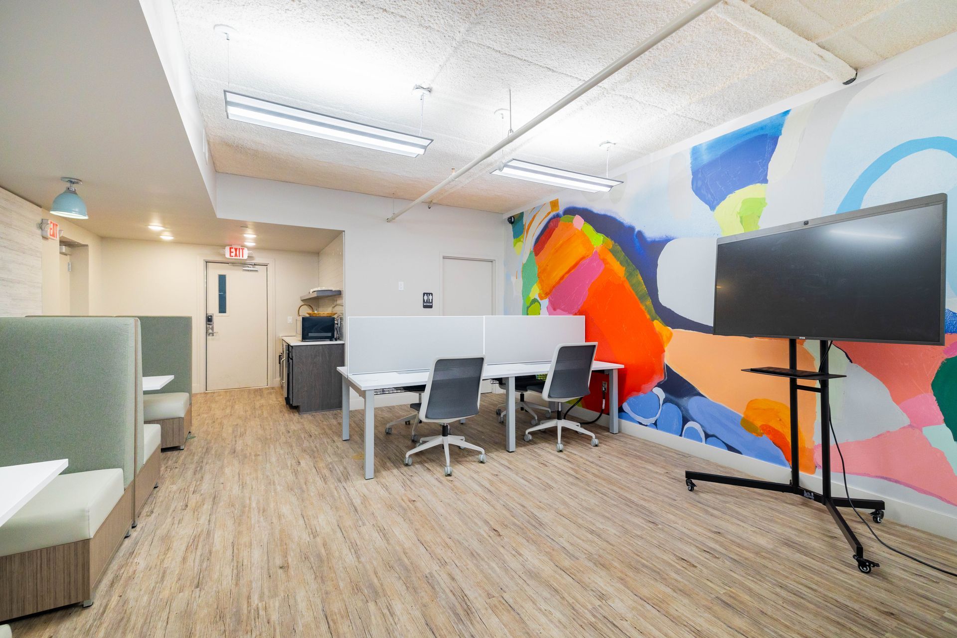 Modern office meeting area with white desks, black chairs, a large TV, and a colorful mural wall