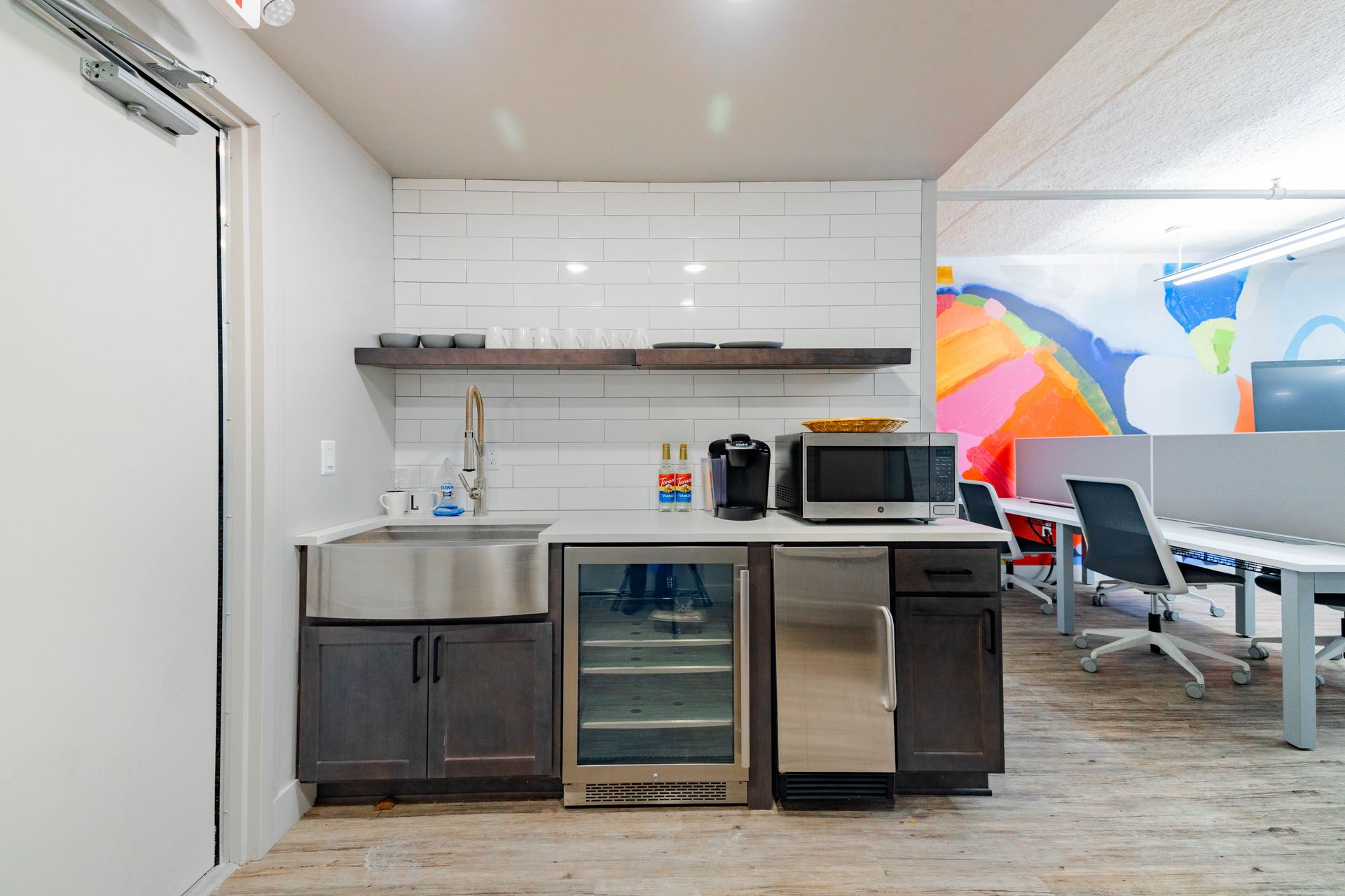 Kitchenette with sink, microwave, mini fridge, and open shelving in a bright room next to tables.