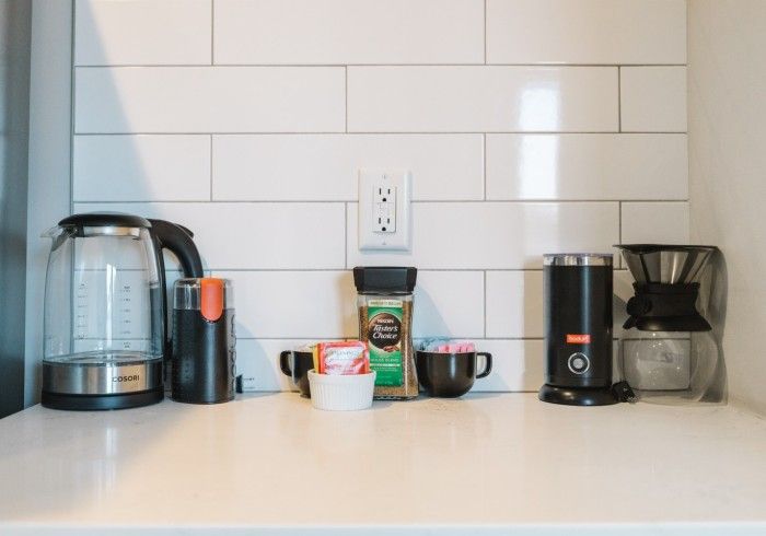 Coffee station: electric kettle, coffee maker, frother, coffee, and mugs on a white countertop.