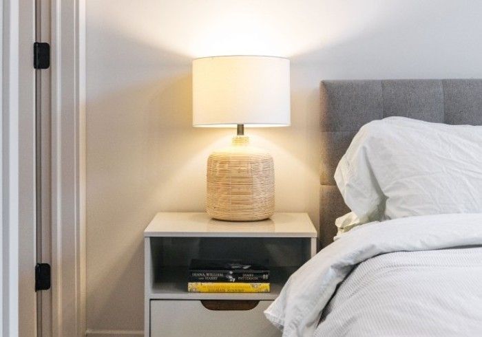 Bedside table with lamp, lit, next to a bed with white bedding, in a room.