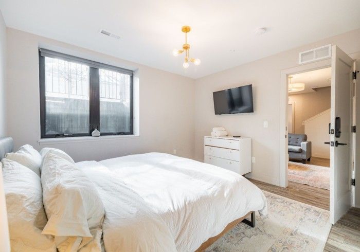 Bedroom with white bedding, TV, dresser, window, open doorway, and gold light fixture.